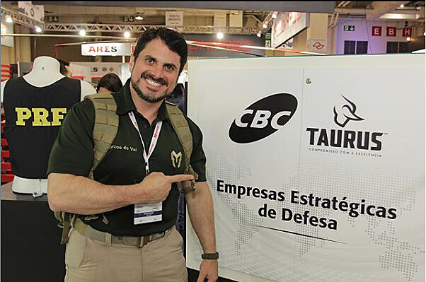Senador, que trabalhava como instrutor, posa ao lado de banner em feira de armas realizada no Brasil