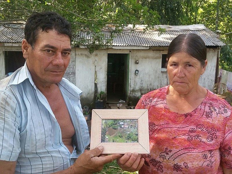 Agricultores Nelson e Eva exibem fotografia do terreno onde viveram por 23 anos. Ao fundo, o casebre para onde se mudaram na sexta-feira