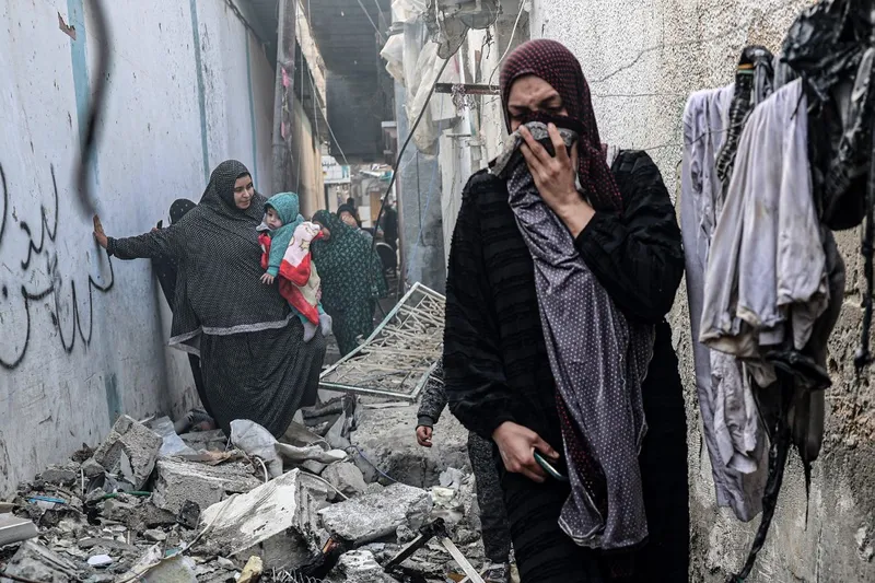 Mulheres caminham sob destroços após bombardeio israelense em Rafah, no sul da Faixa de Gaza, no domingo (25)