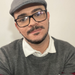 Autorretrato do colunista. Um homem cis branco, da bigode e barba pretas, usa óculos e uma boina cinza.