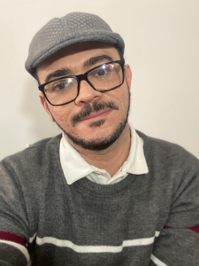 Autorretrato do colunista. Um homem cis branco, da bigode e barba pretas, usa óculos e uma boina cinza.
