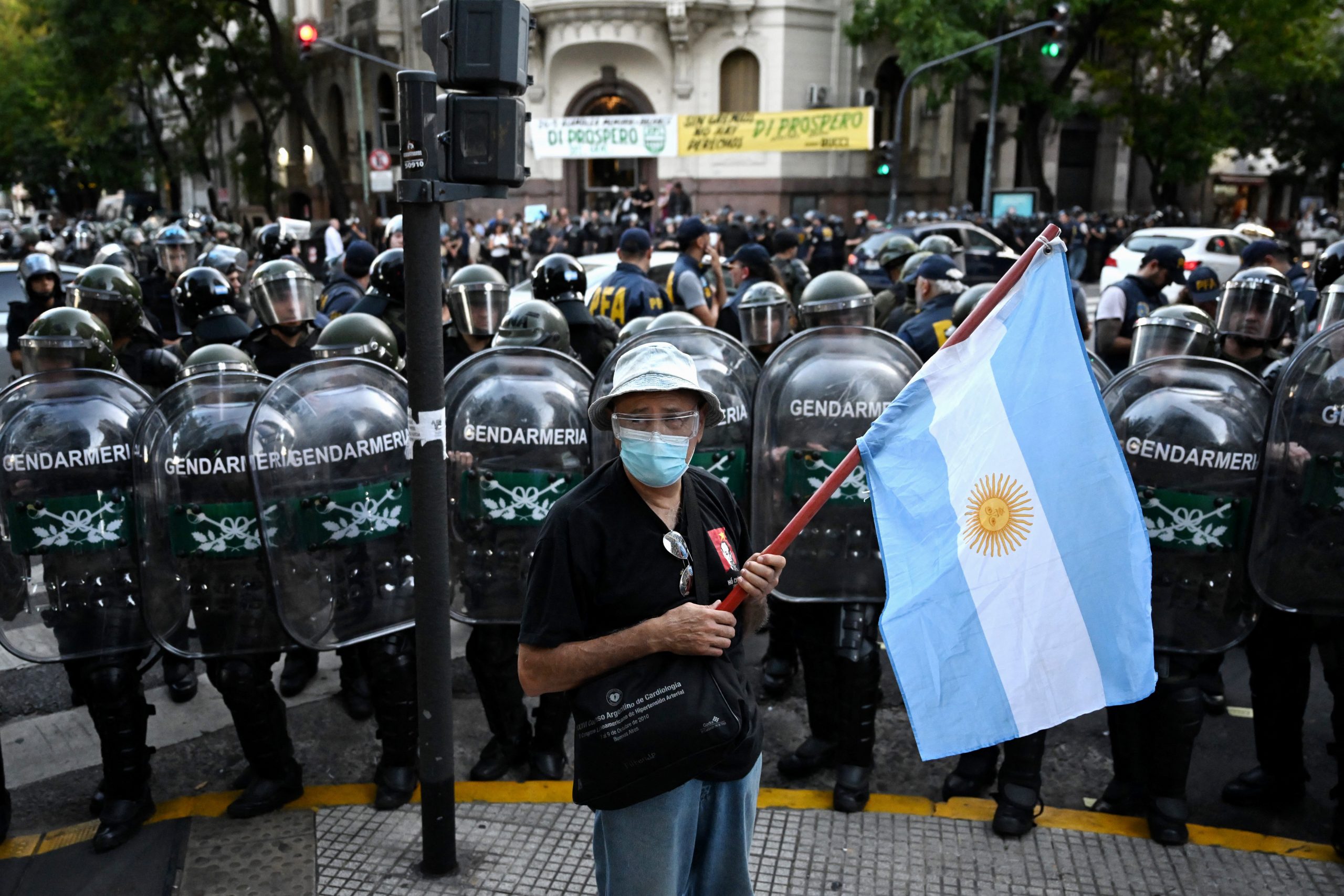 Mulher idosa segura a bandeira da Argentina. Atrás, uma barreira de policiais a cercam