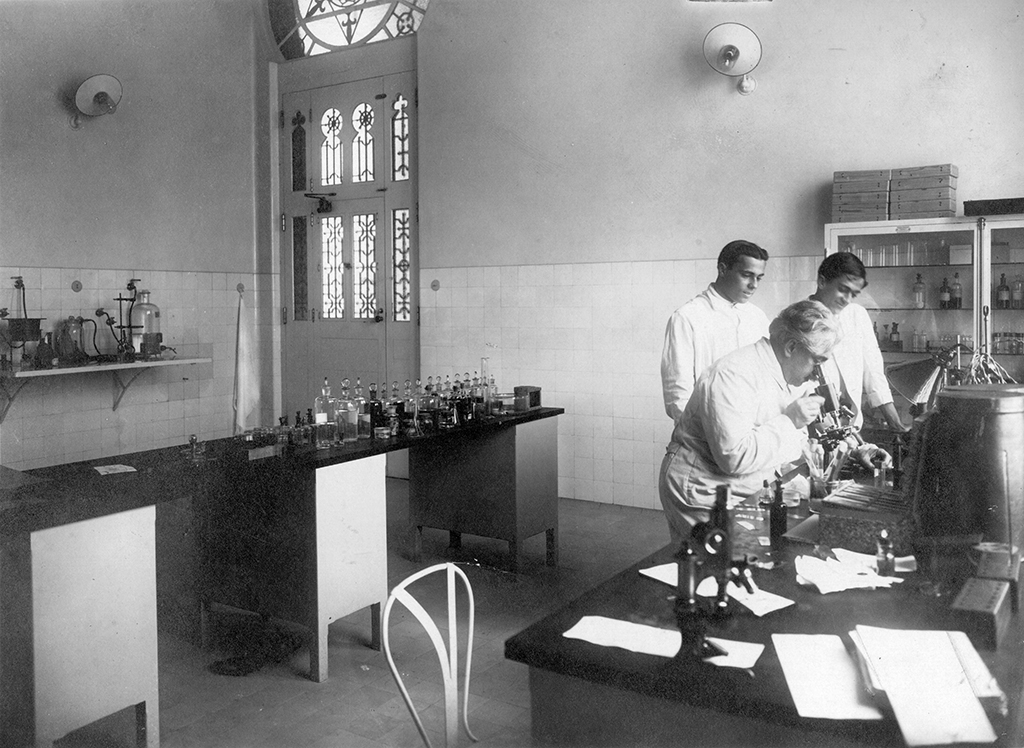 Oswaldo Cruz no laboratório de Manguinhos, observado por seu filho Bento Oswaldo Cruz e pelo pesquisador Burle de Figueiredo