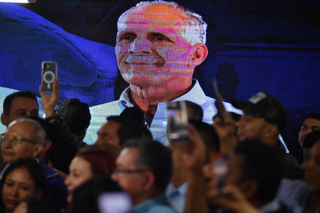 Uma tela de LCD exibe um retrato do candidato presidencial de Honduras, Nasry Asfura, do Partido Nacional, enquanto apoiadores celebram após o Conselho Nacional Eleitoral (CNE) declará-lo vencedor da eleição presidencial em Tegucigalpa, em 24 de dezembro de 2025.
