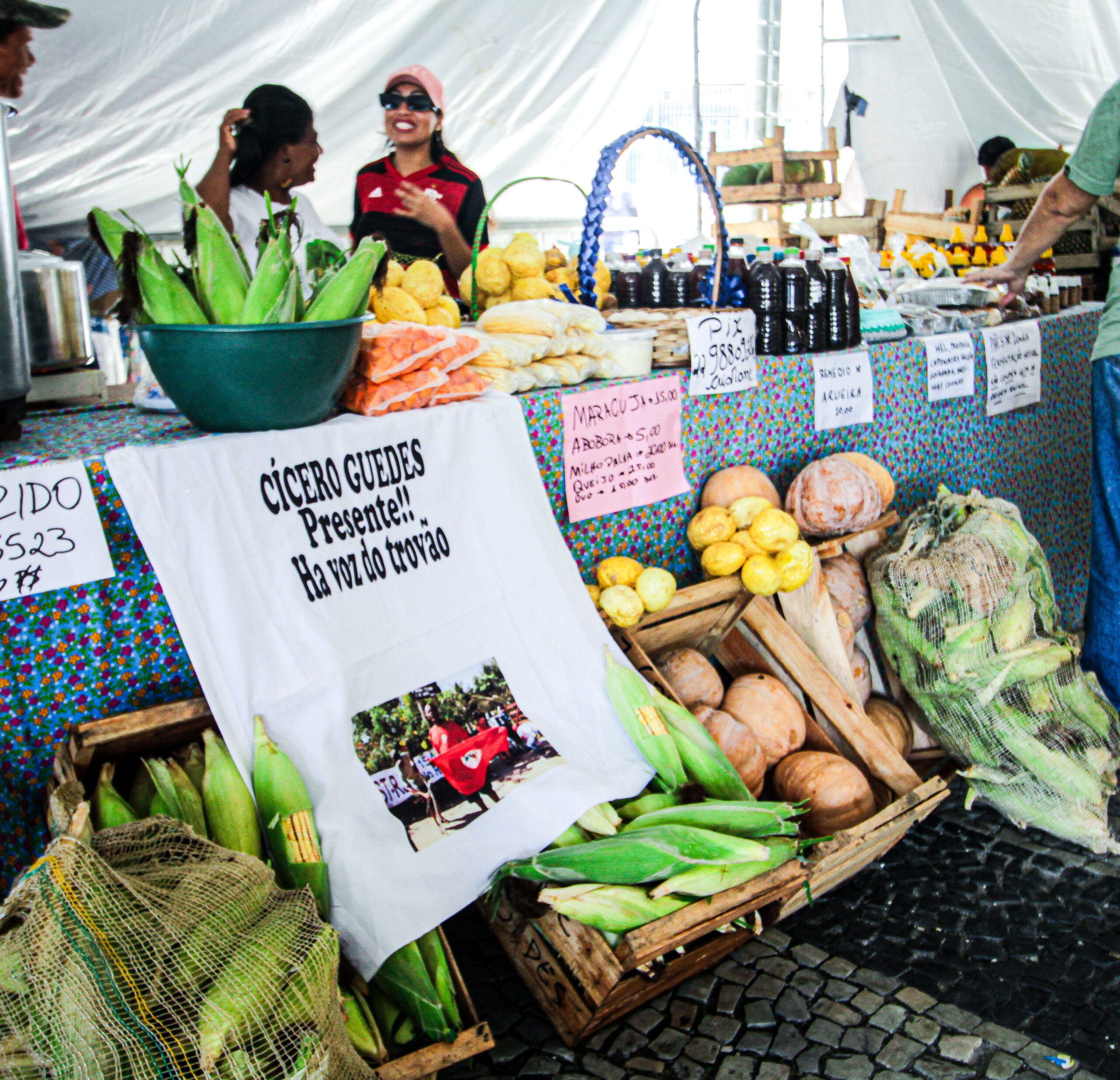 Participantes da 15ª edição da Feira expõe seus produtos como mel, verduras e frutas. Ao fundo, duas mulheres, uma delas vestindo a camisa do Flamengo, conversam