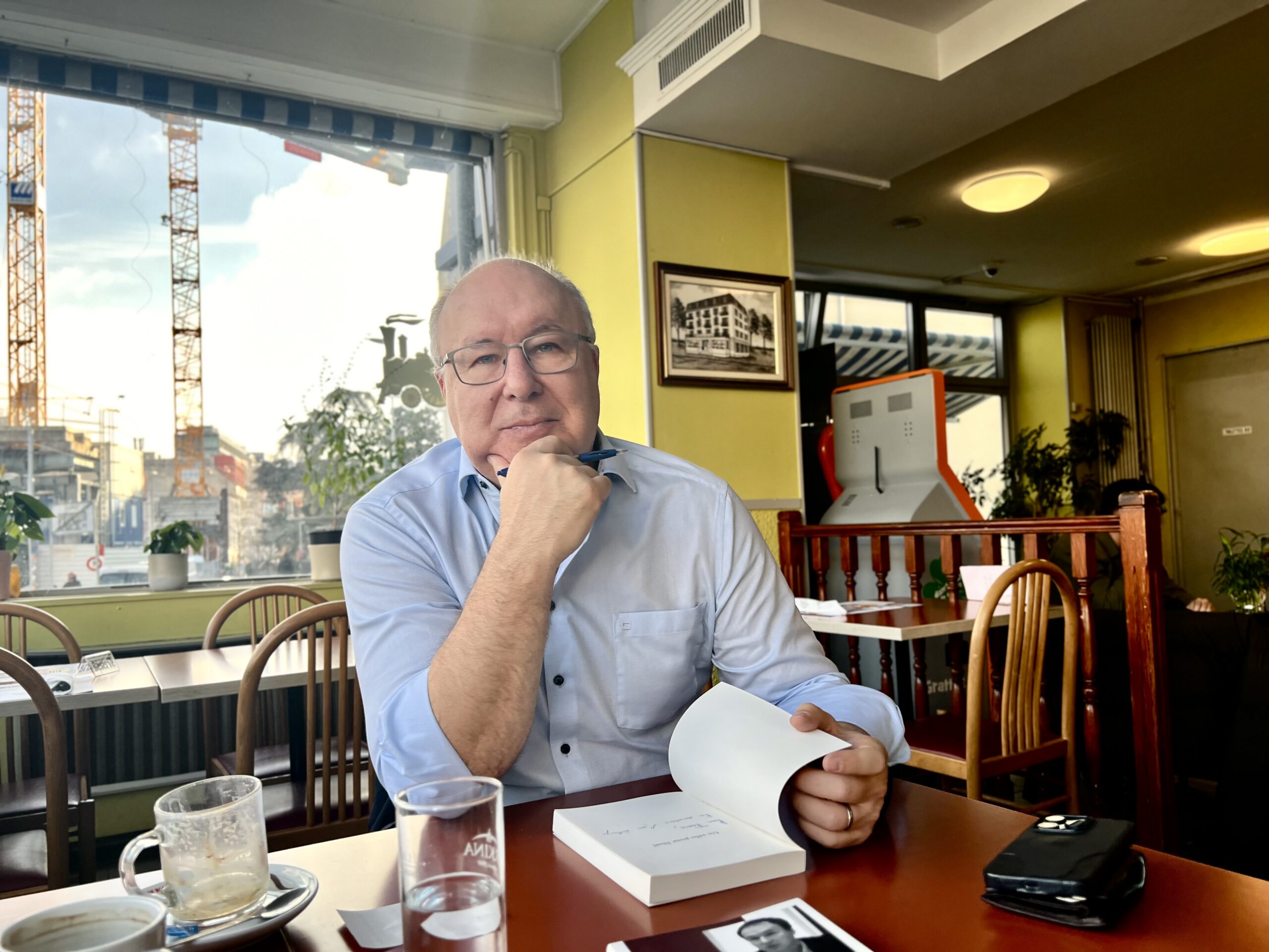 Pierre-Yves Maillard é um homem branco, careca e usa óculos. Ele está sentado em um bistrô em Rennes, interior da suiça, e apoia um dos cotovelos na mesa e segura o queixo com a mão direita. Com a outra, abre um livro