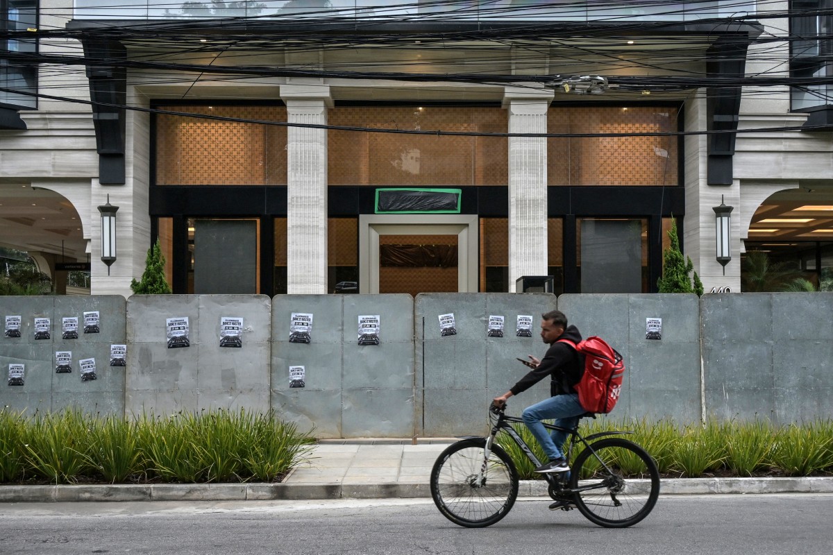 A man rides his bike past the facade of the Banco Master protected by hoardings, with the bank's logo covered in plastic, in Sao Paulo, Brazil, on January 22, 2026, after Brazil's Central Bank ordered its shutdown due to a major liquidity crunch and "serious violations" of financial regulations. (Photo by NELSON ALMEIDA / AFP)