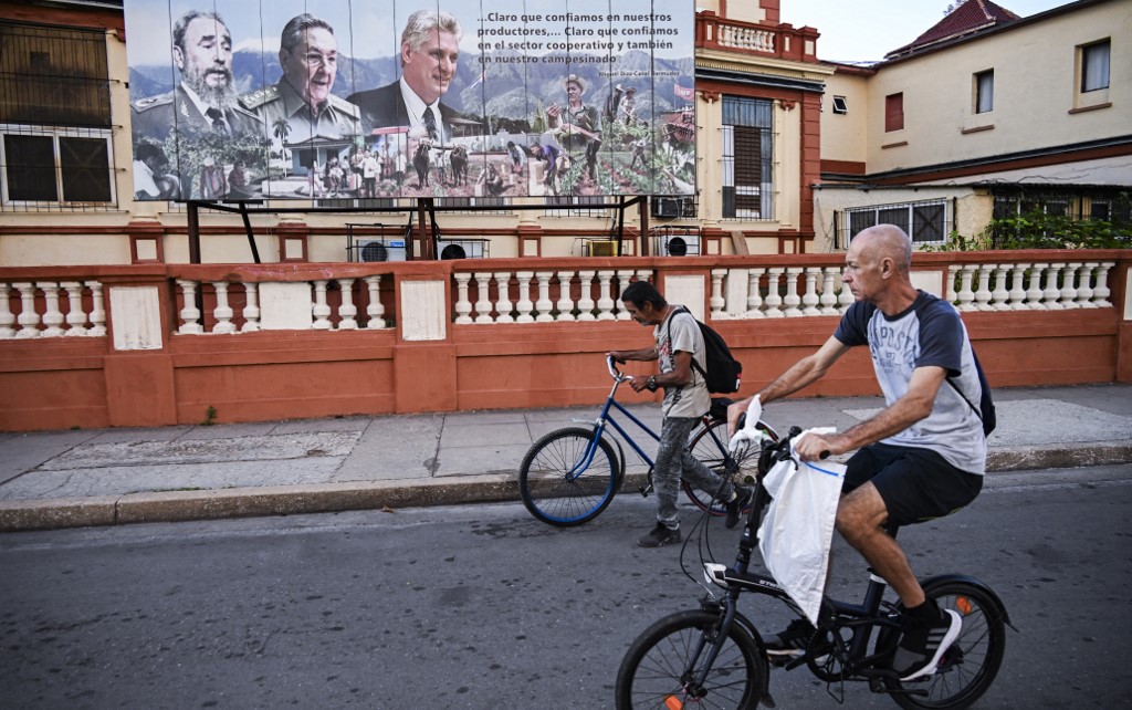 Dois homens passam de bicicleta por um outdoor que retrata o falecido líder cubano Fidel Castro, o ex-presidente Raúl Castro e o presidente Miguel Díaz-Canel, em Havana, em 16 de fevereiro de 2026.