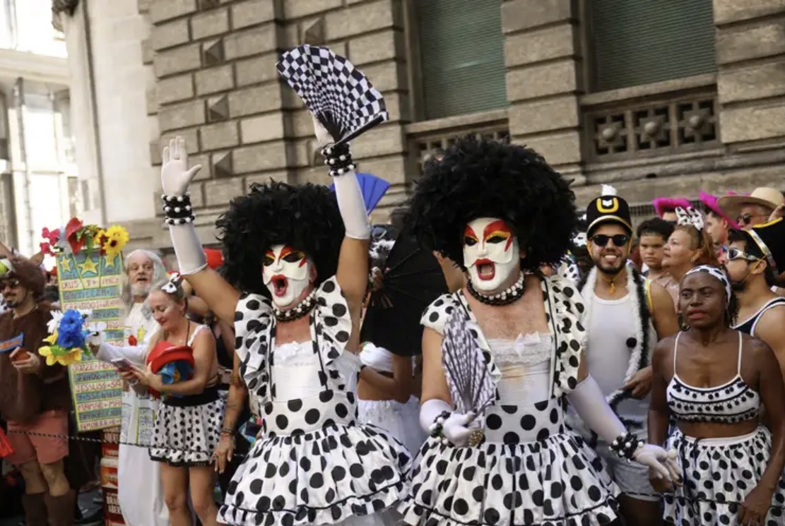 A imagem mostra dois homens vestidos com roupas femininas, de vestido na cor branca com bolinhas pretas, peruca preta e rostos pintados de branco, cores da identidade visual do bloco Cordão do Bola Preta, durante apresentação do bloco em rua no Centro do Rio