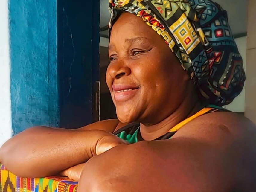Tia Cida é uma mulher negra que veste um touca em tecido africano e está com os braços cruzados e apoiados em uma divisória de madeira que leva a cozinha do Quilombo Ferreira Diniz e está decorada com um tecido africano