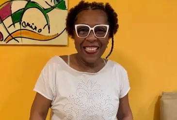 Andrea Marins, mulher negra de cabelos curtos, usa óculos com borda de acetato na cor branca e veste uma blusa branca lisa. Ela posa para foto e ao fundo há uma parede amarelo mostarda e um quadro com pintura abstrata.