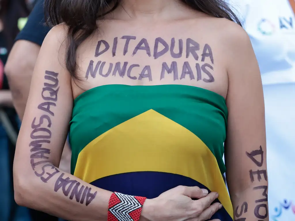 Mulher usa bandeira do Brasil como blusa e em seu colo é possível ler Ditadura nunca mais