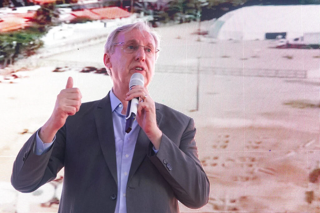 Celso Pansera é um homem alto, branco, de cabelos brancos e segura um microfone em cerimônia de abertura para apresentá-lo ao cargo de presidente da Codemar. Ao fundo está projetada uma imagem de obras.