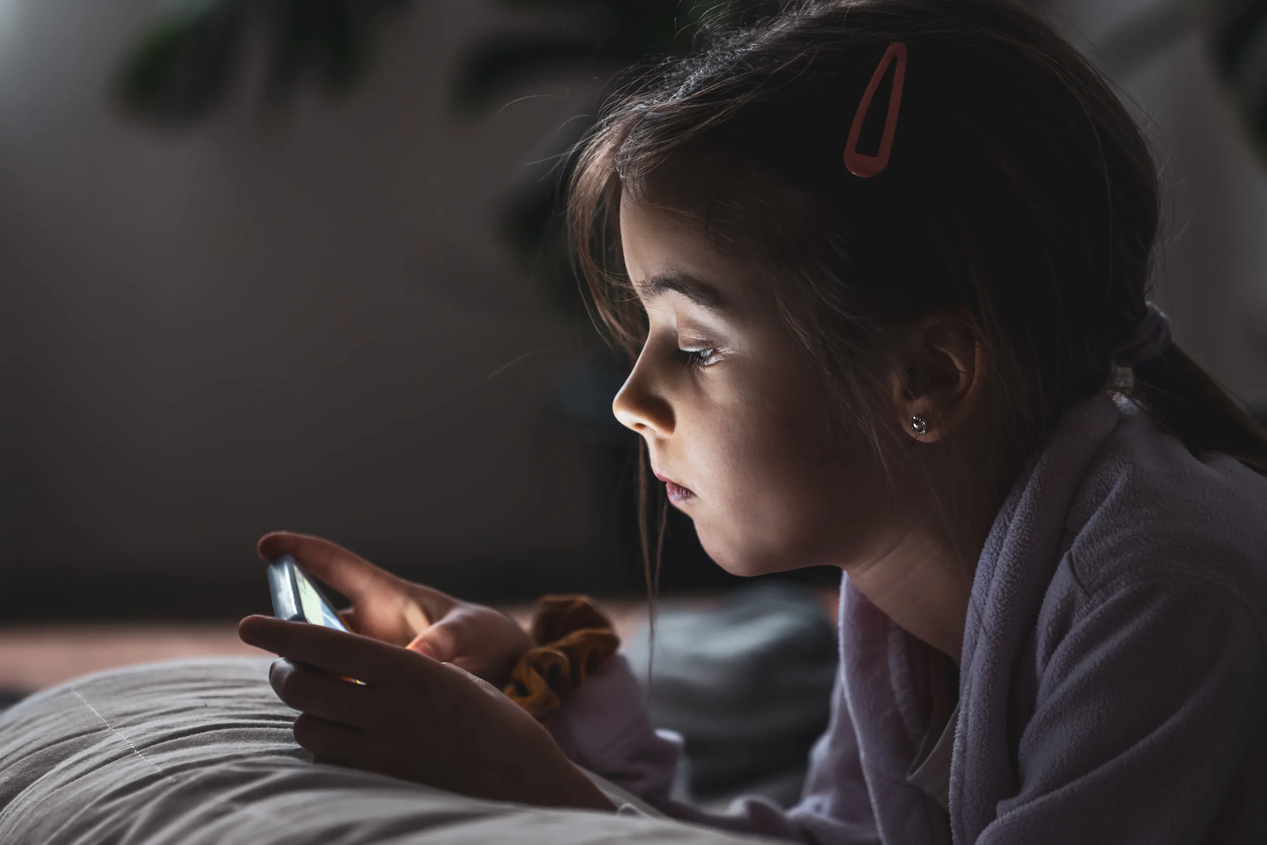 Little girl uses smartphone, lying on pillow at home, gadgets addiction concept.