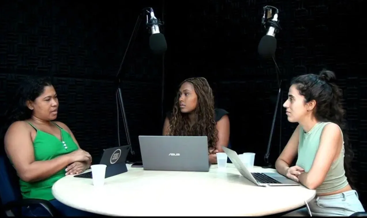 Na foto, três mulheres estão sentadas em volta de uma mesa branca redonda para a gravação do podcast