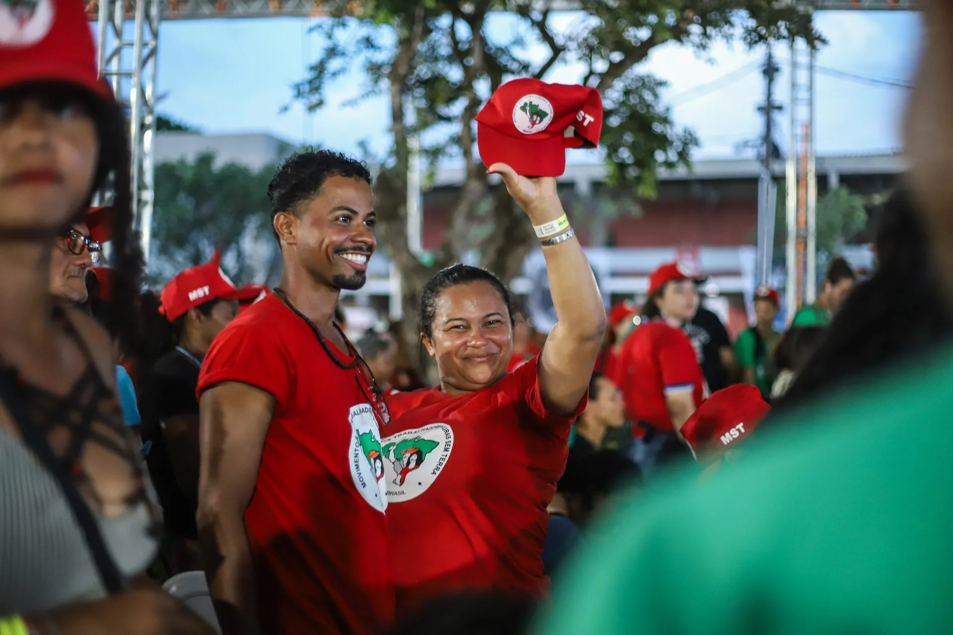 Em encontro Nacional do MST, um companheiro e uma companheira posam lado a lado com a camisa do MST. Com sua mão esquerda ela levanta o boné do Movimento dos Trabalhadores Sem Terra