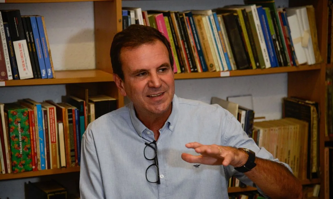 Eduardo Paes registrado em entrevista. Homem branco, cabelos curtos, com relógio no pulso, veste camisa social azul clara. Ao fundo uma estante de madeira com vários livros