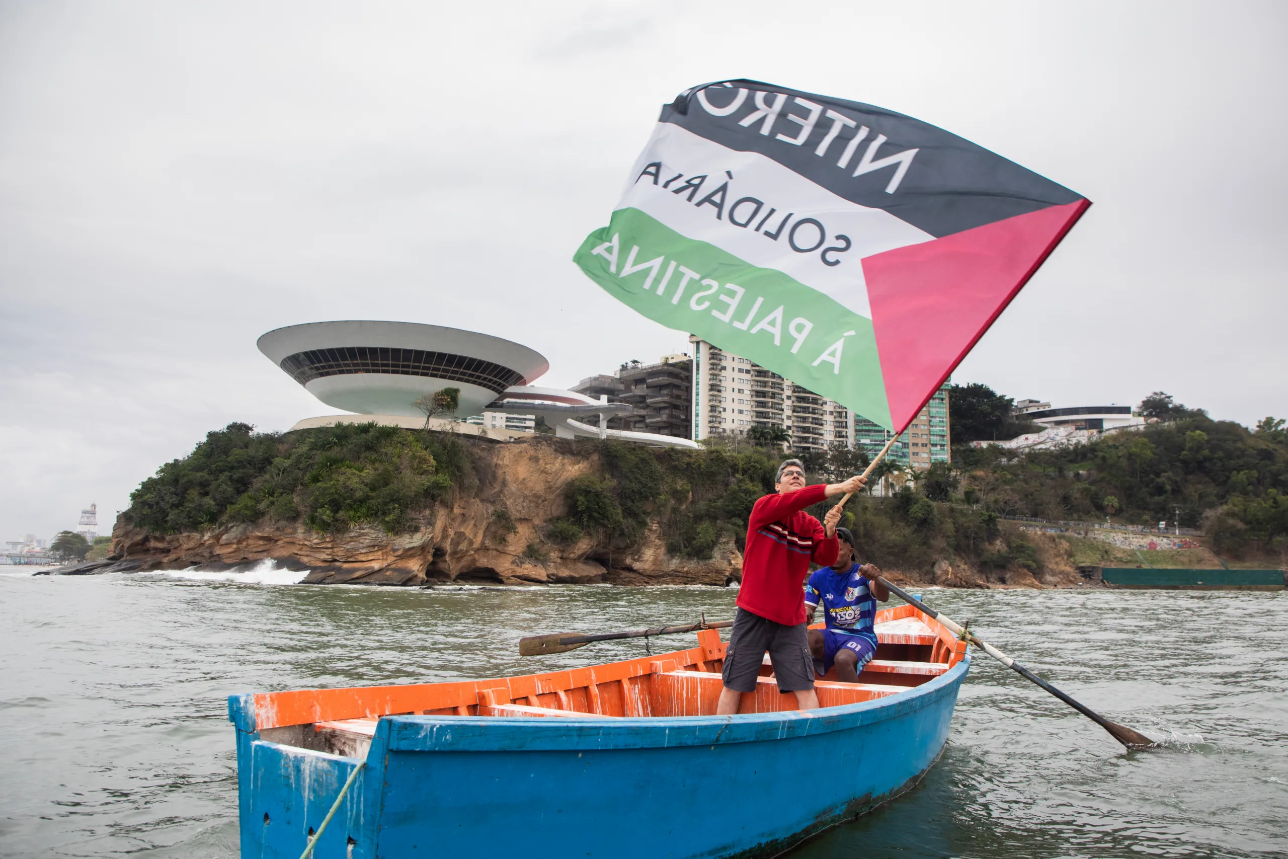 Manifestante em barco na praia de Icaraí, em Niterói, carrega bandeira de Palestina. Ao fundo está o Museu Niemeyer