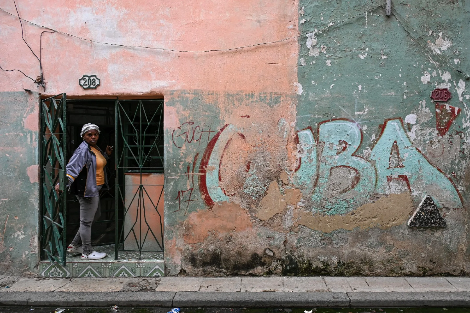 Uma mulher espreita pela porta de sua casa ao lado de um muro com um grafite escrito "CUBA" em uma rua de Havana, Cuba, em 6 de maio de 2026.