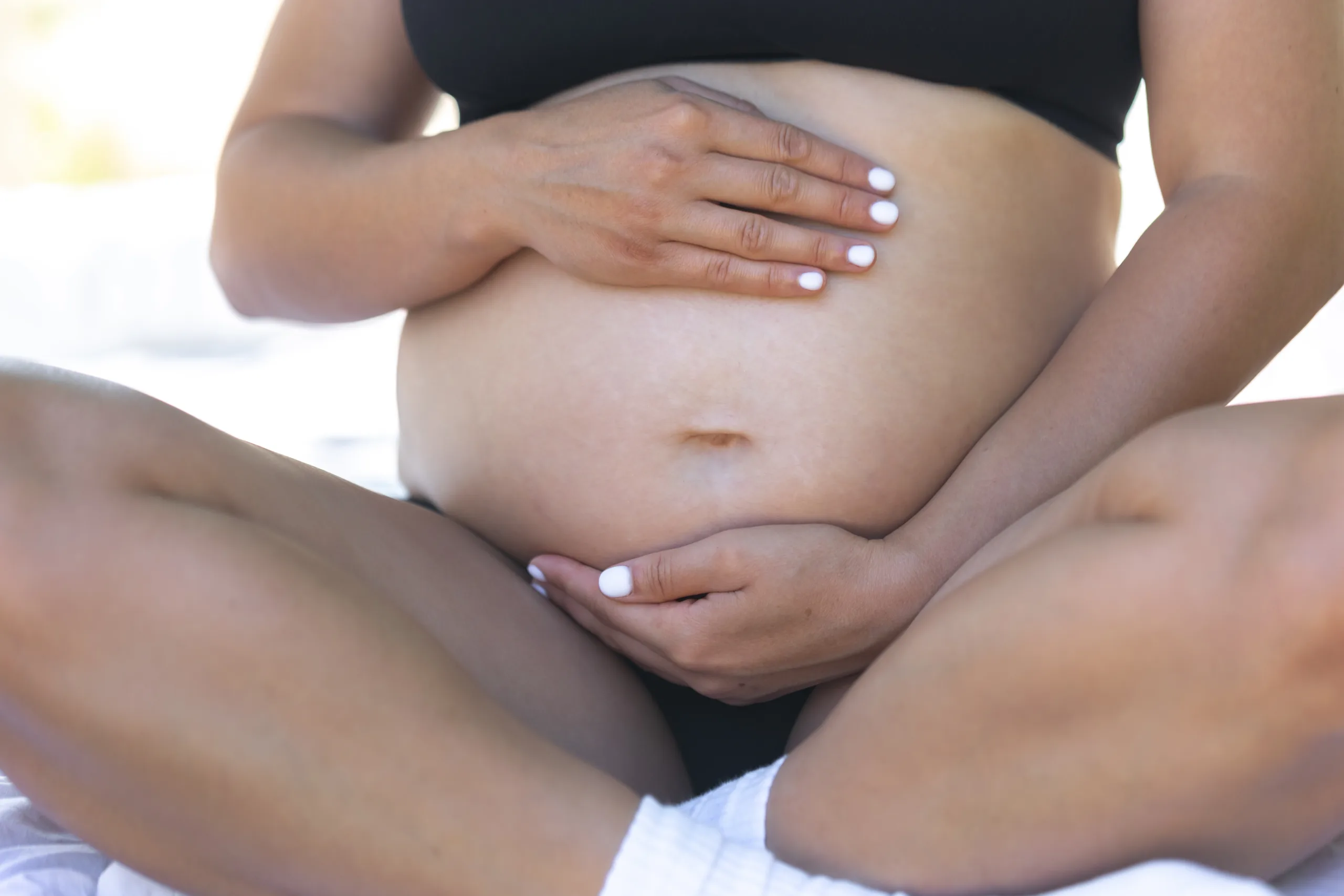 Foto registra gravidez de mulher negra anônima e mostra ela sentada em uma cama, de pernas cruzadas e colocando as mãos em cima de barriga, como se a segurasse