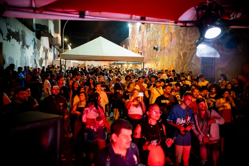 Multidão reunida em um evento cultural noturno ao ar livre, em uma rua cercada por grandes murais artísticos. Sarau-vá