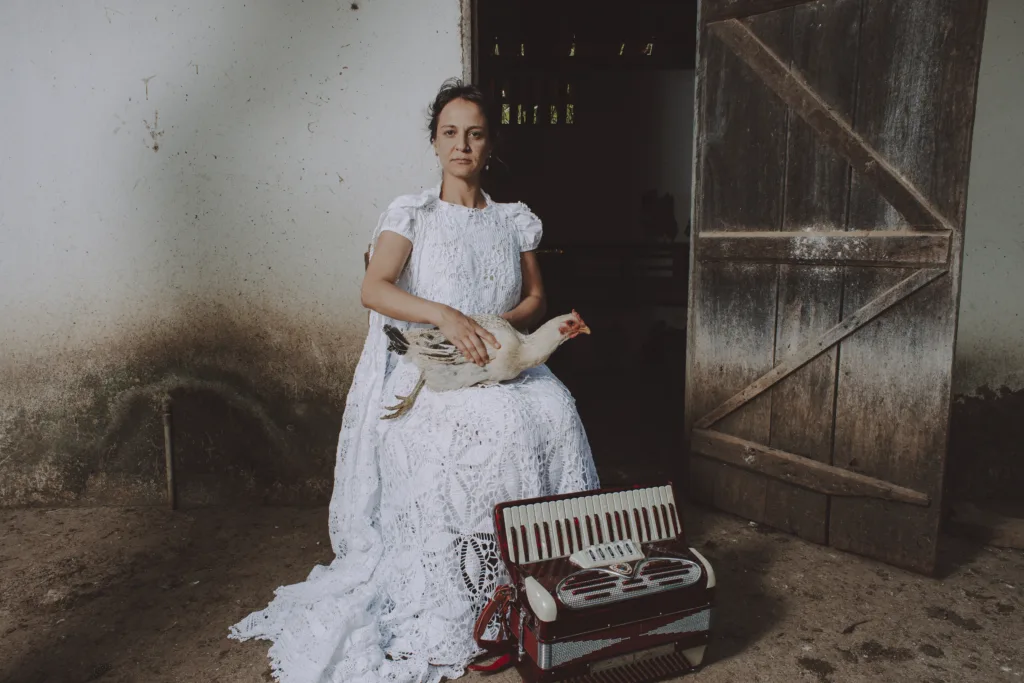 Vestida de branco, mulher segura uma galinha enquanto posa ao lado de uma sanfona na entrada de uma casa rural.