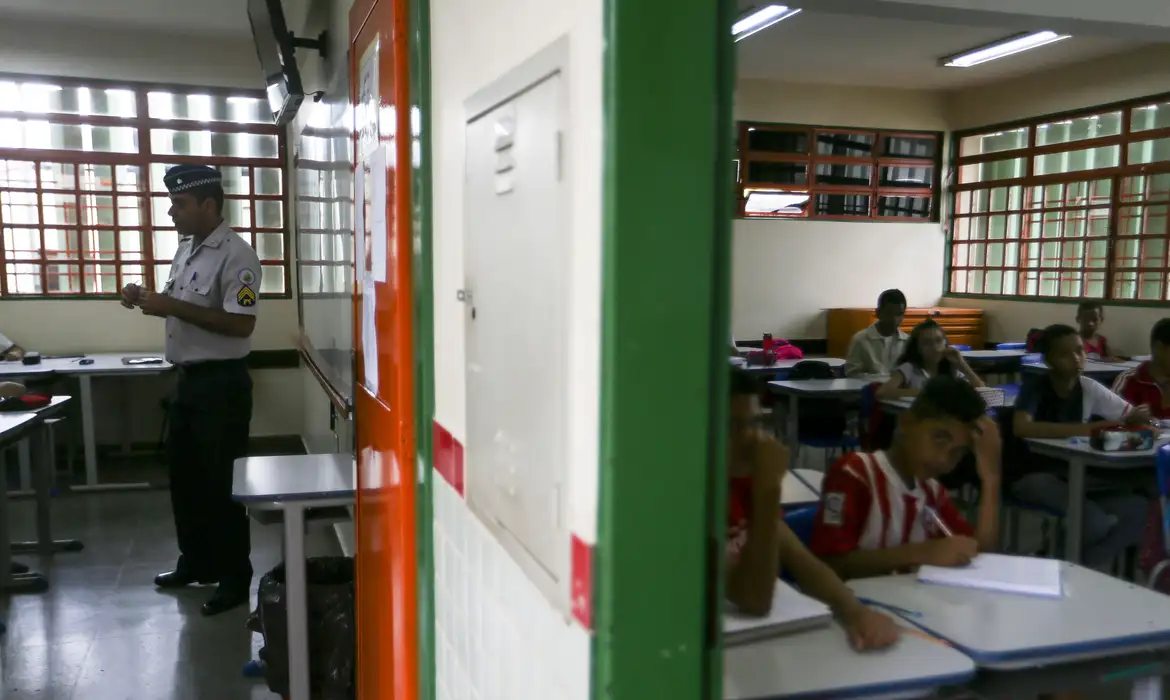 Fotografia mostra um policial militar falando para crianças em uma sala de aula