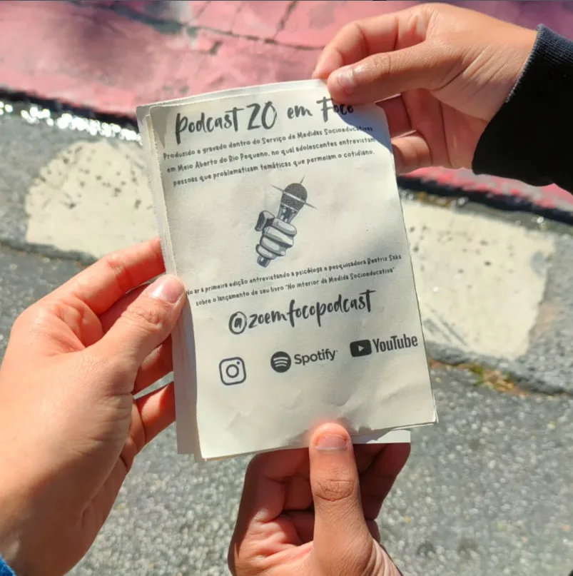 As mãos de três jovens seguram um panfleto do podcast 'ZO em Foco'