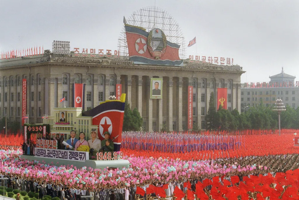 A portrait of North Korean President Kim Il-sung hangs from the facade of an official building in Pyongyang 09 September 1988 while a float to the glory of the regime passes through a huge crowd during a monster parade celebrating the secretive Stalinist nation's 40th anniversary. Kim Il-sung (1912-94) founded the Korean People's Revolutionary Army in 1932, and led a long struggle against the Japanese. He proclaimed the Republic in 1948 and became effective head of state, establishing a unique personality cult and a Stalinist political-economic system. Kim named his son, Kim Jong-il as his successor. After his death, Kim Il-sung was named "eternal president" of the Democratic People's Republic of Korea. (Photo by ROBERTO COLOMA / AFP)