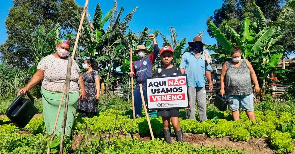 Uma criança segura um cartaz com o texto "agrotóxico mata". Ao redor, estão adultos agricultores. Estão todos em um lugar que aparenta ser uma horta
