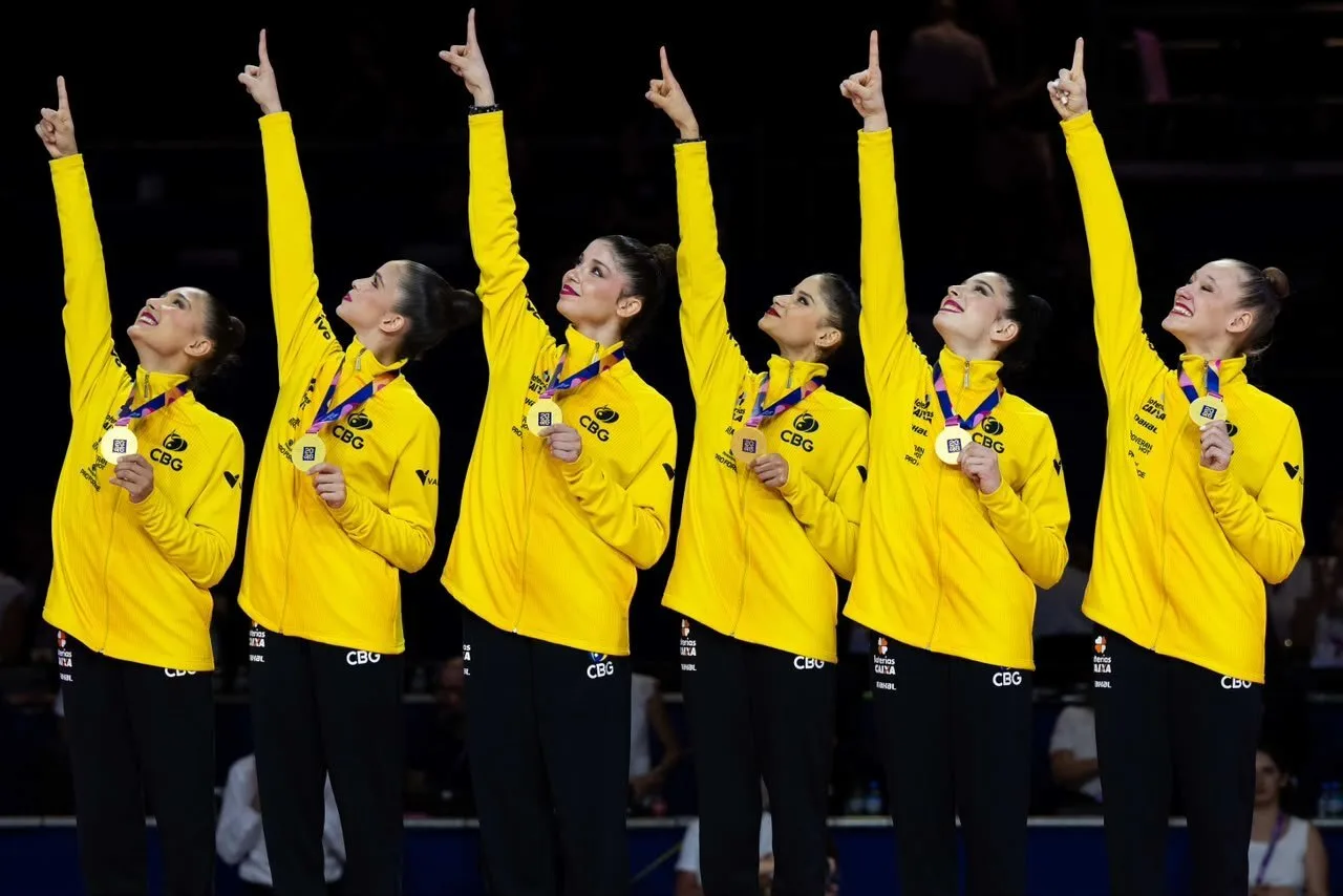 Atletas da equipe brasileira de ginástica rítimica pousam para foto oficial, uma ao lado da outra, todas apontando e olhando para o lado superior direito delas e segurando com a mão esquerda a medalha de ouro presa ao pescoço de cada uma. Estão vestidas com blusa de moletom amarelo com símbolo da seleção brasileira no peito esquerdo.