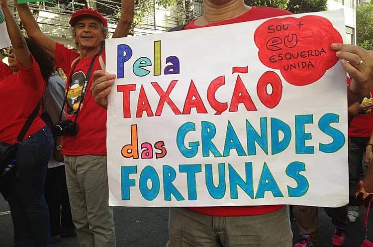 Em um protesto, uma pessoa segura um cartaz com o texto "pela taxação das grandes fortunas"