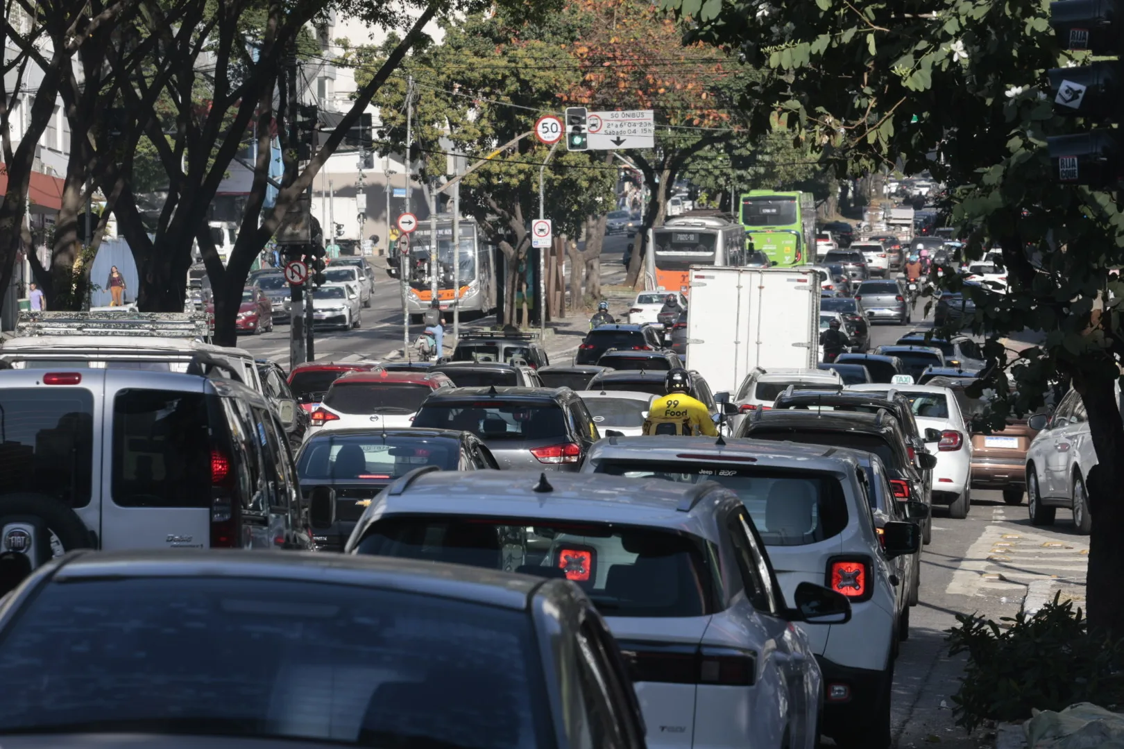 Trânsito intenso em avenida urbana, com carros, ônibus e motocicletas ocupando as vias
