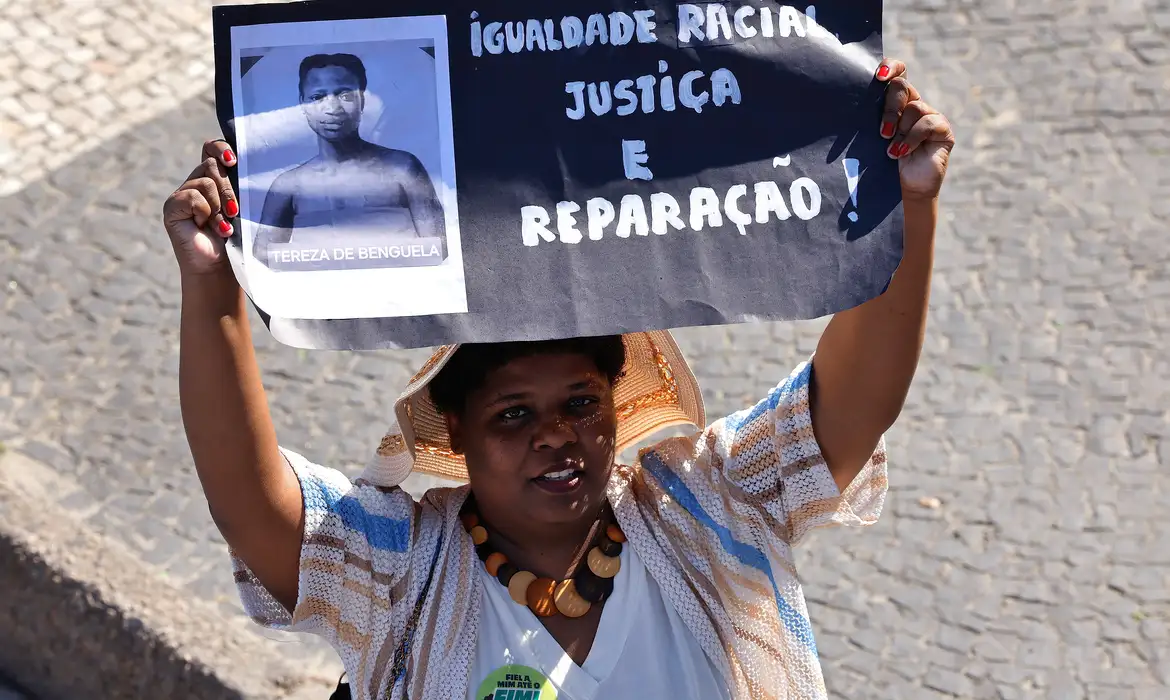 Mulher negra segura cartaz onde se lê 'Igualdade racial, justiça e reparação'