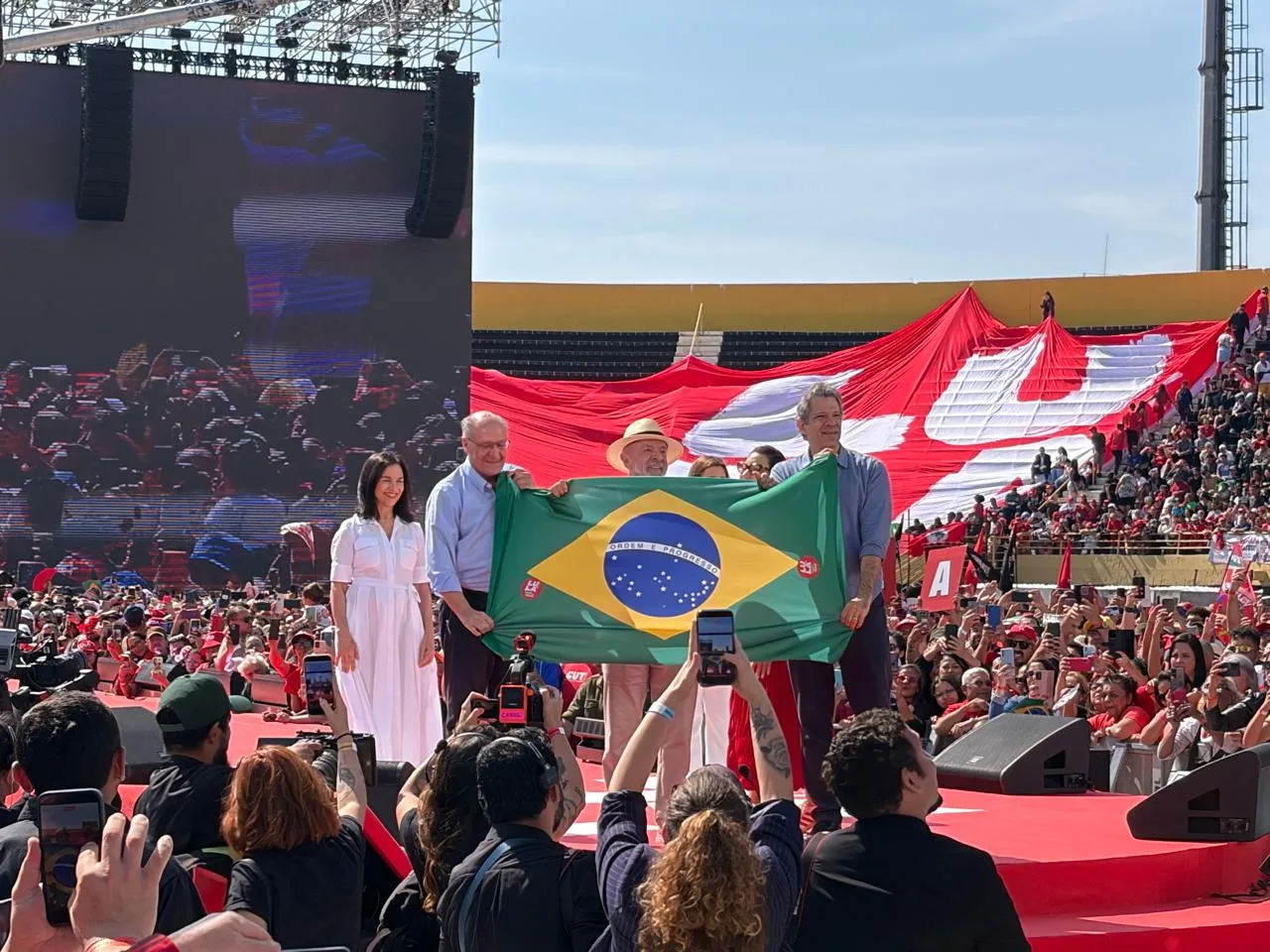 Lu Alckimin, Geraldo Alckimin, Lula, Janja e Haddad estão sobre um palco em estádio segurando uma bandeira do Brasil, enquanto o público está por atrás, segurando bandeira com o nome de Lula.
