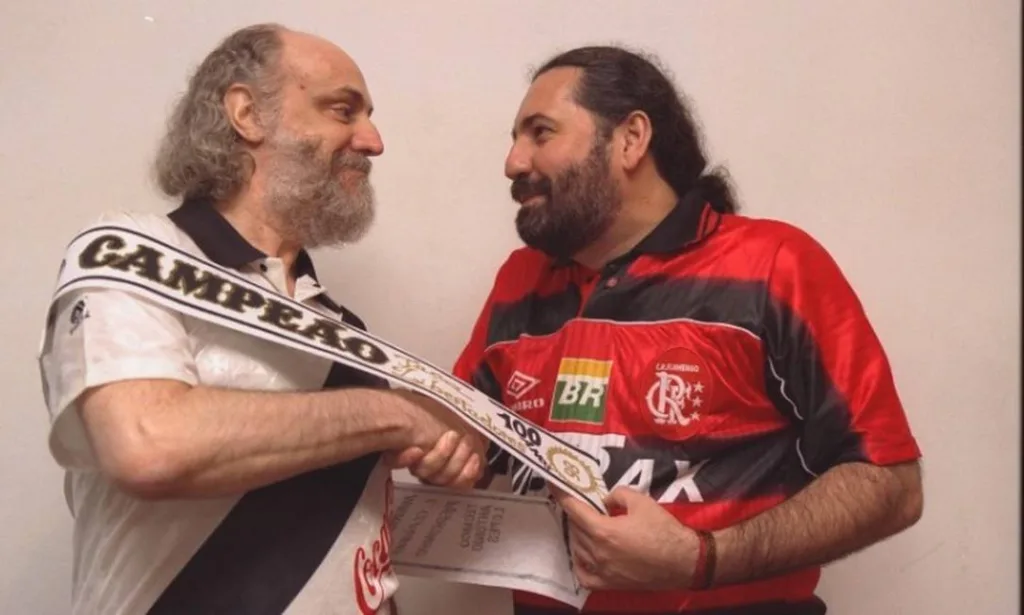 Fotografia de Aldir Blac, com uma camiseta do Vasco da Gama, apertando a mão de Moacyr Luz, que usa camiseta do Flamento