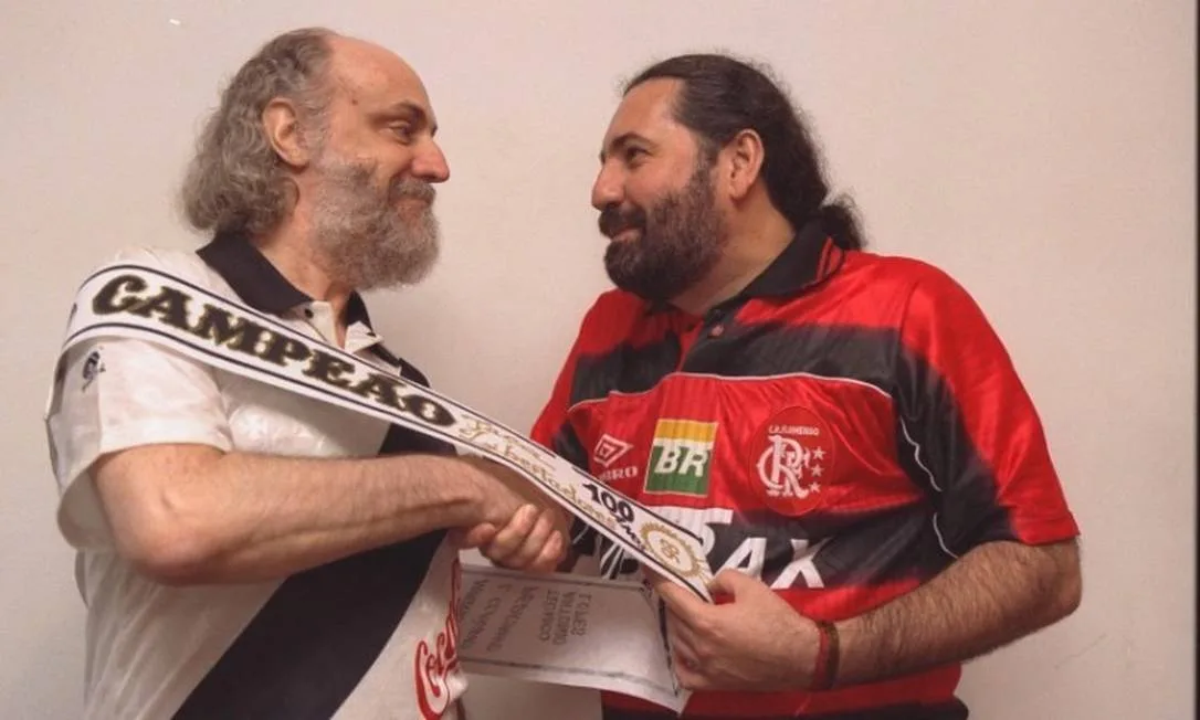 Fotografia de Aldir Blac, com uma camiseta do Vasco da Gama, apertando a mão de Moacyr Luz, que usa camiseta do Flamento