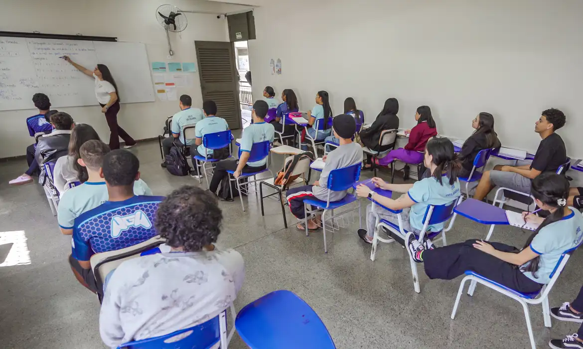 Fotografia de uma sala de aula com vários adolescentes de costas em carteiras