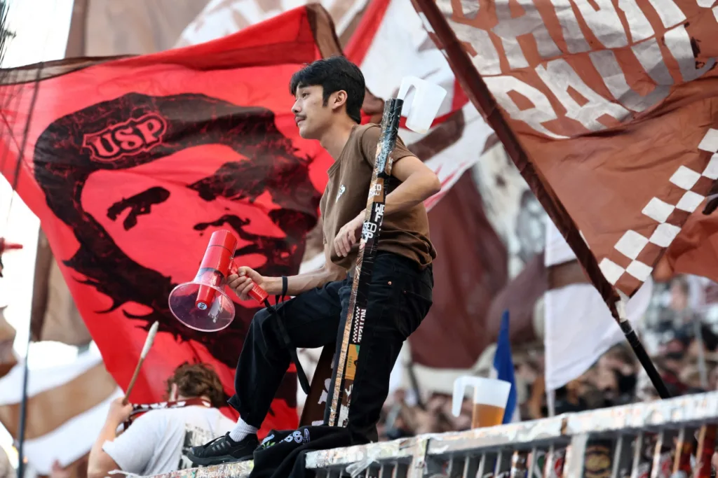 Um torcedor do St. Pauli está sobre uma barreira ao lado de uma bandeira com a imagem de Che Guevara durante a partida entre FC St. Pauli e Borussia Dortmund, válida pela primeira divisão do Campeonato Alemão (Bundesliga), em Hamburgo, no norte da Alemanha, em 23 de agosto de 2025.