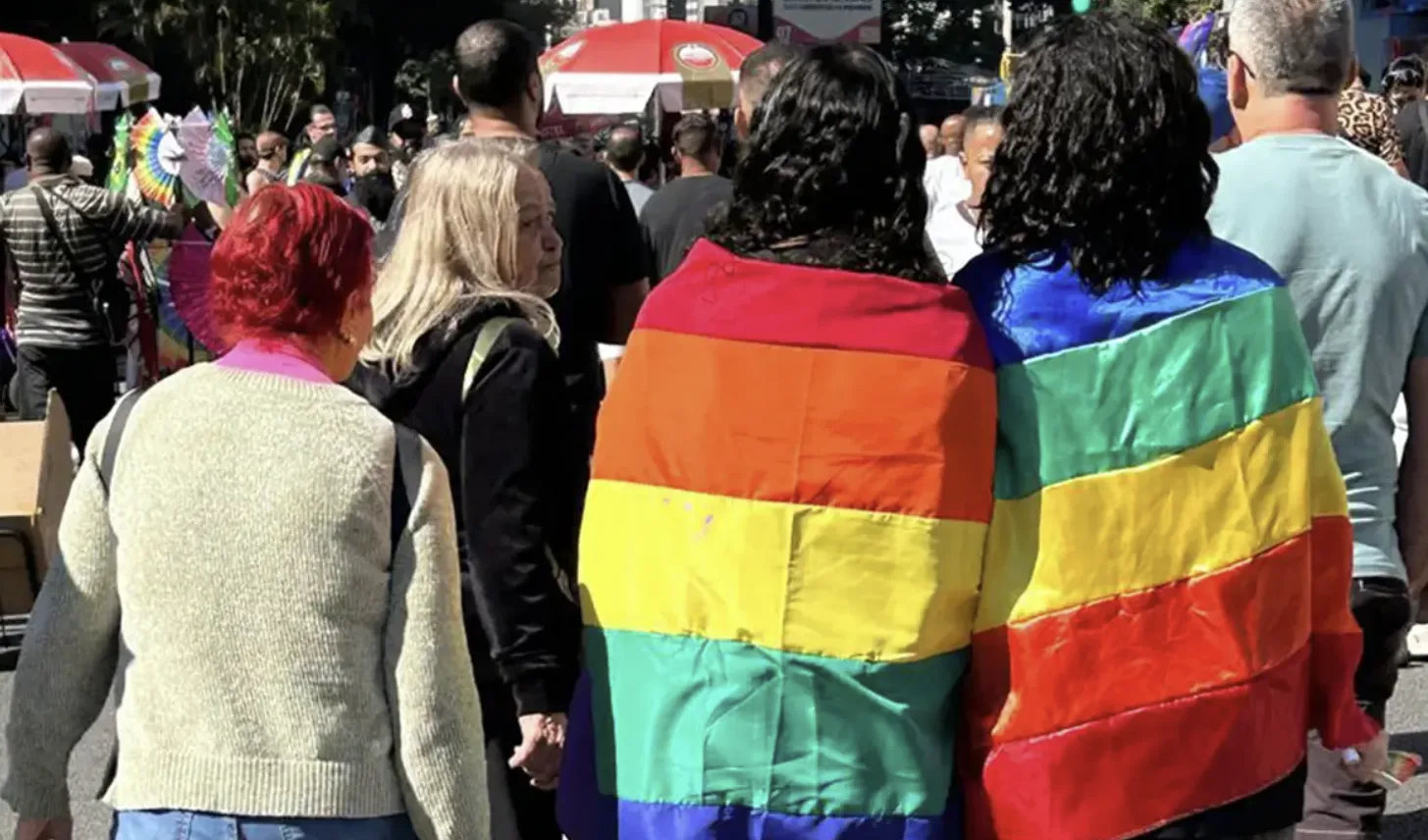 Imagem mostra três mulheres de costa, cobertas por bandeira com as cores do arco-íris, que simboliza a luta LGBTQIAPN+