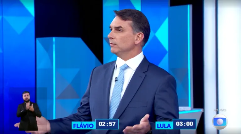 Flávio está no estúdio da Globo, durante debate de segundo turno entre Lula e Flávio. Ele está vestido de terno e gravata azuis. É um homem branco, com cabelo curto, na faixa etária de 45 anos de idade.