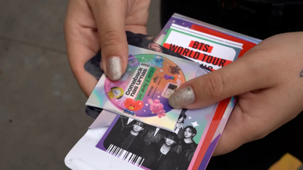 Fotografia de mãos segurando um ingresso para o show do BTS e um adesivo com o texto "comeback nas urnas".